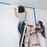 Essential DIY Skills That Save Money as Costs Keep Rising A couple working together to paint a wall, one on a ladder applying tape and the other observing