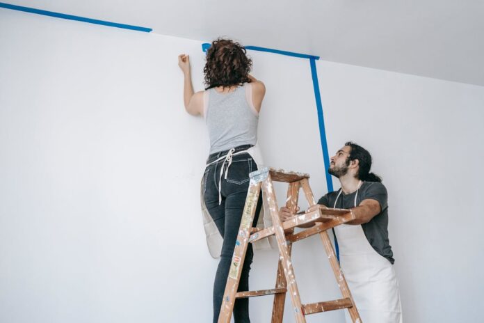 A couple working together to paint a wall, one on a ladder applying tape and the other observing