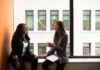 Why Companies Are Pushing Mindfulness Instead of Fixing Burnout Two women in business attire engaged in a friendly conversation by a window