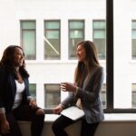 Why Companies Are Pushing Mindfulness Instead of Fixing Burnout Two women in business attire engaged in a friendly conversation by a window