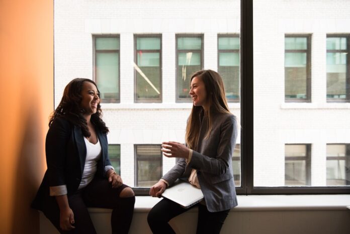 Two women in business attire engaged in a friendly conversation by a window
