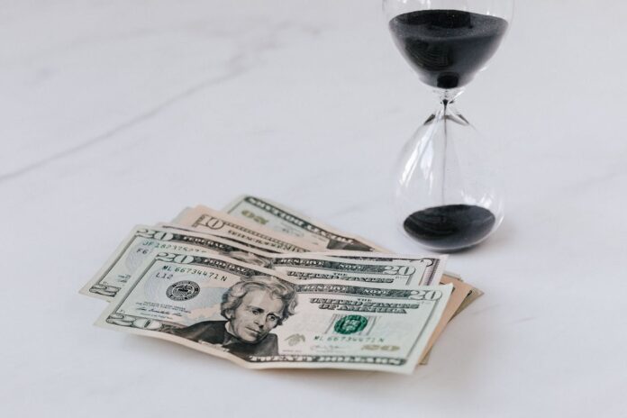 A stack of dollar bills next to an hourglass filled with black sand