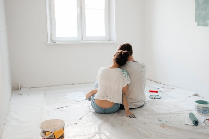 pexels-maria-ovchinnikova-46787598-7509790 (1).jpg A couple sitting on the floor of a freshly painted room, surrounded by painting supplies