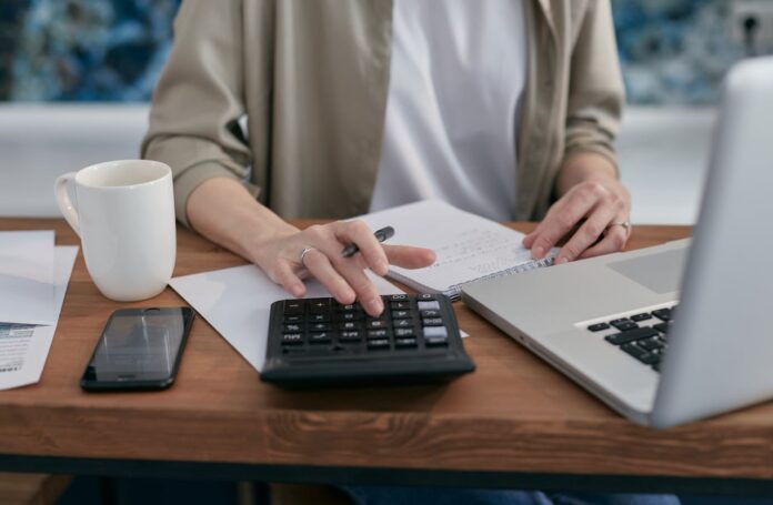 Person using a calculator while working at a desk with a laptop and notebook